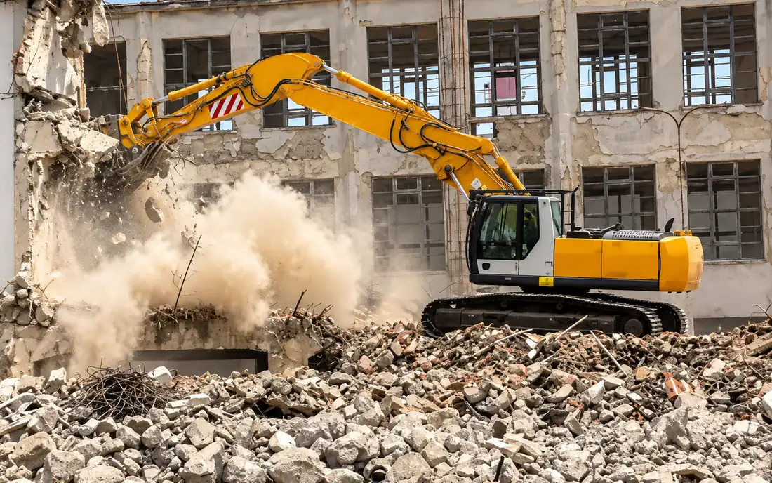 Excavator with a hydraulic hammer bringing down a concrete building amid dust and rubble