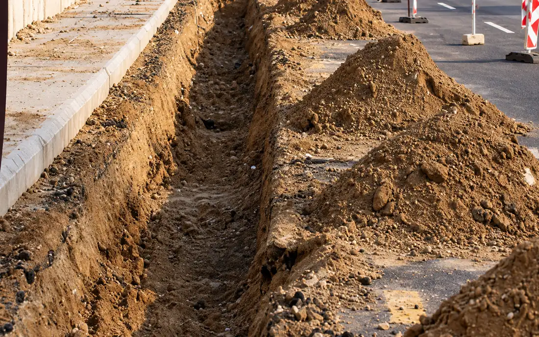 Narrow excavated trench alongside a road curb, with the spoil heaped beside it